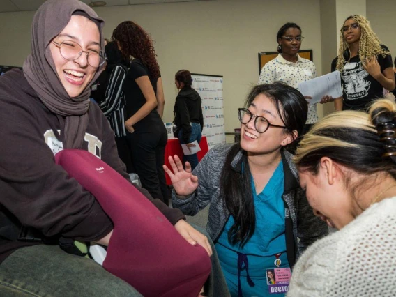 A woman wearing a hijab sits on a chair with two other women, all appearing to share a friendly discussion.