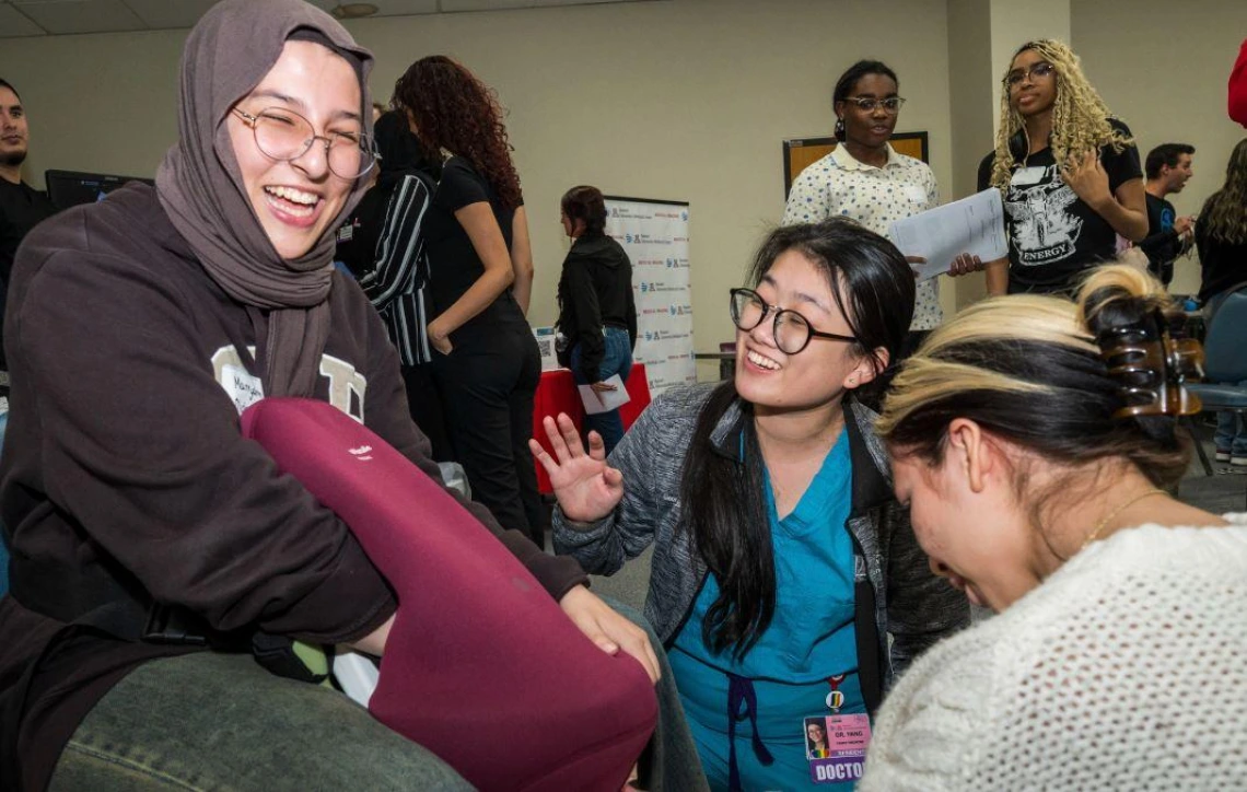 A woman wearing a hijab sits on a chair with two other women, all appearing to share a friendly discussion.